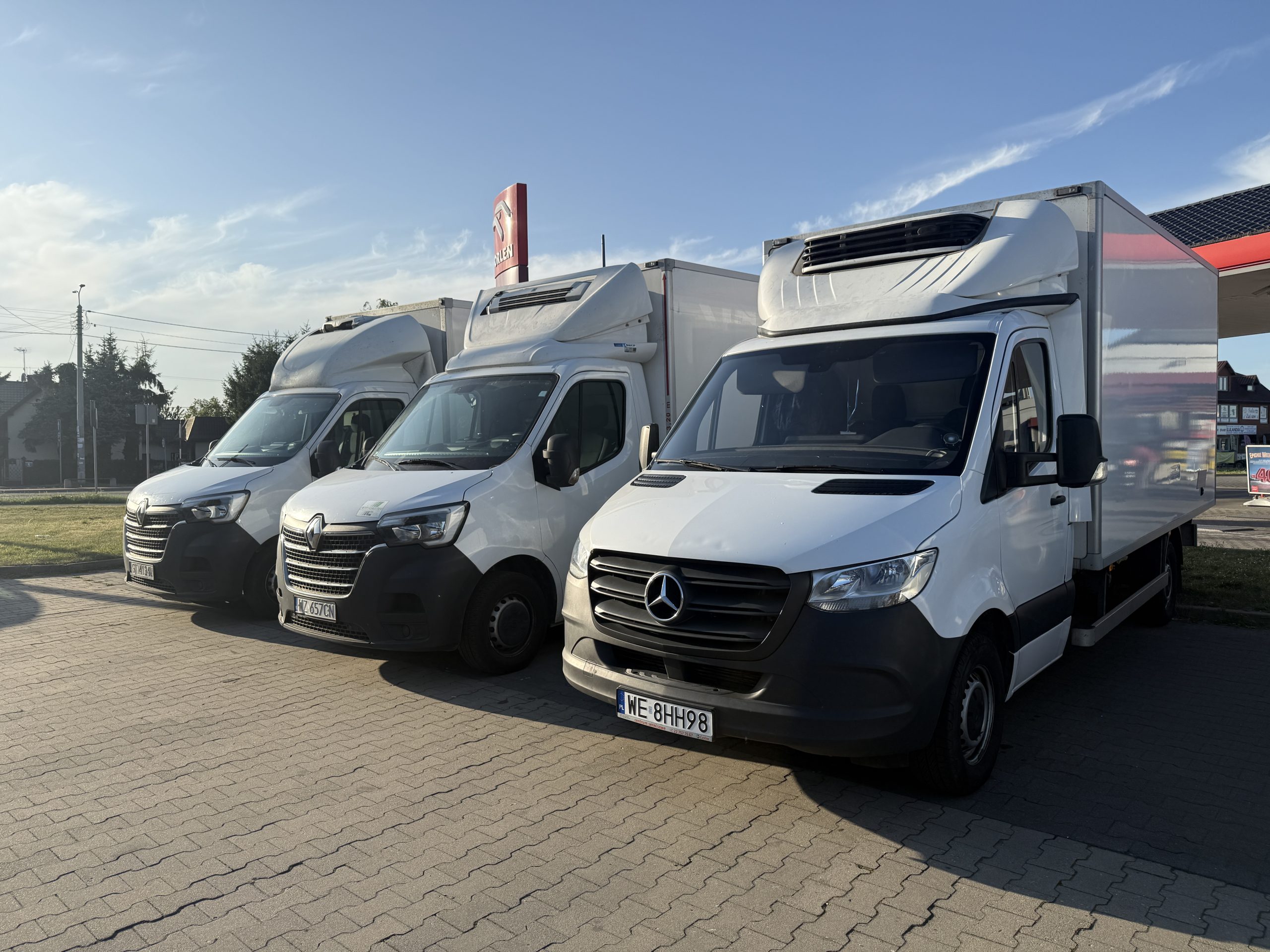 Trzy białe samochody dostawcze z agregatami chłodniczymi, zaparkowane obok siebie na parkingu pod lekkim kątem. Od prawej Mercedes-Benz, pośrodku Renault Master.