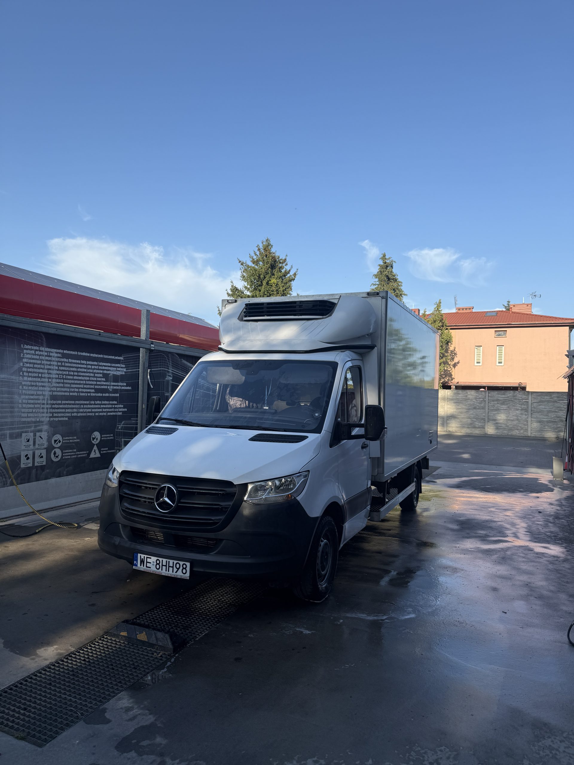 Biały samochód dostawczy Mercedes-Benz z chłodnią stoi na mokrej posadzce myjni samochodowej. W tle widać czerwono-białą konstrukcję myjni i drzewa.